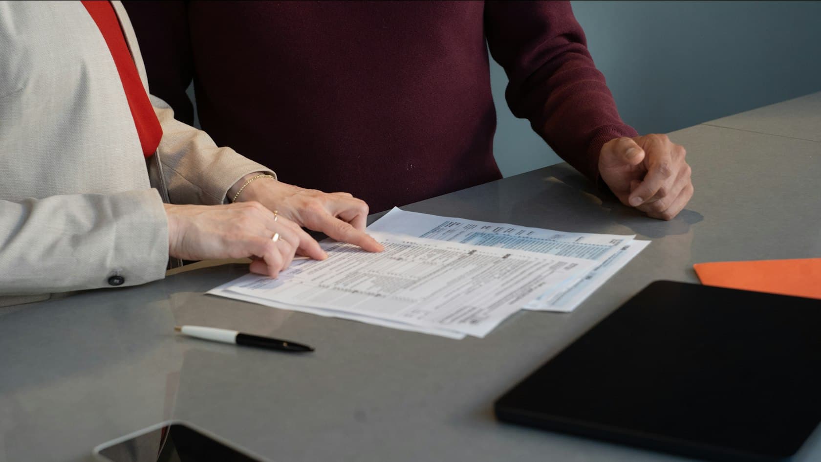 Two colleagues at a table point through printed forms beside a pen and a tablet