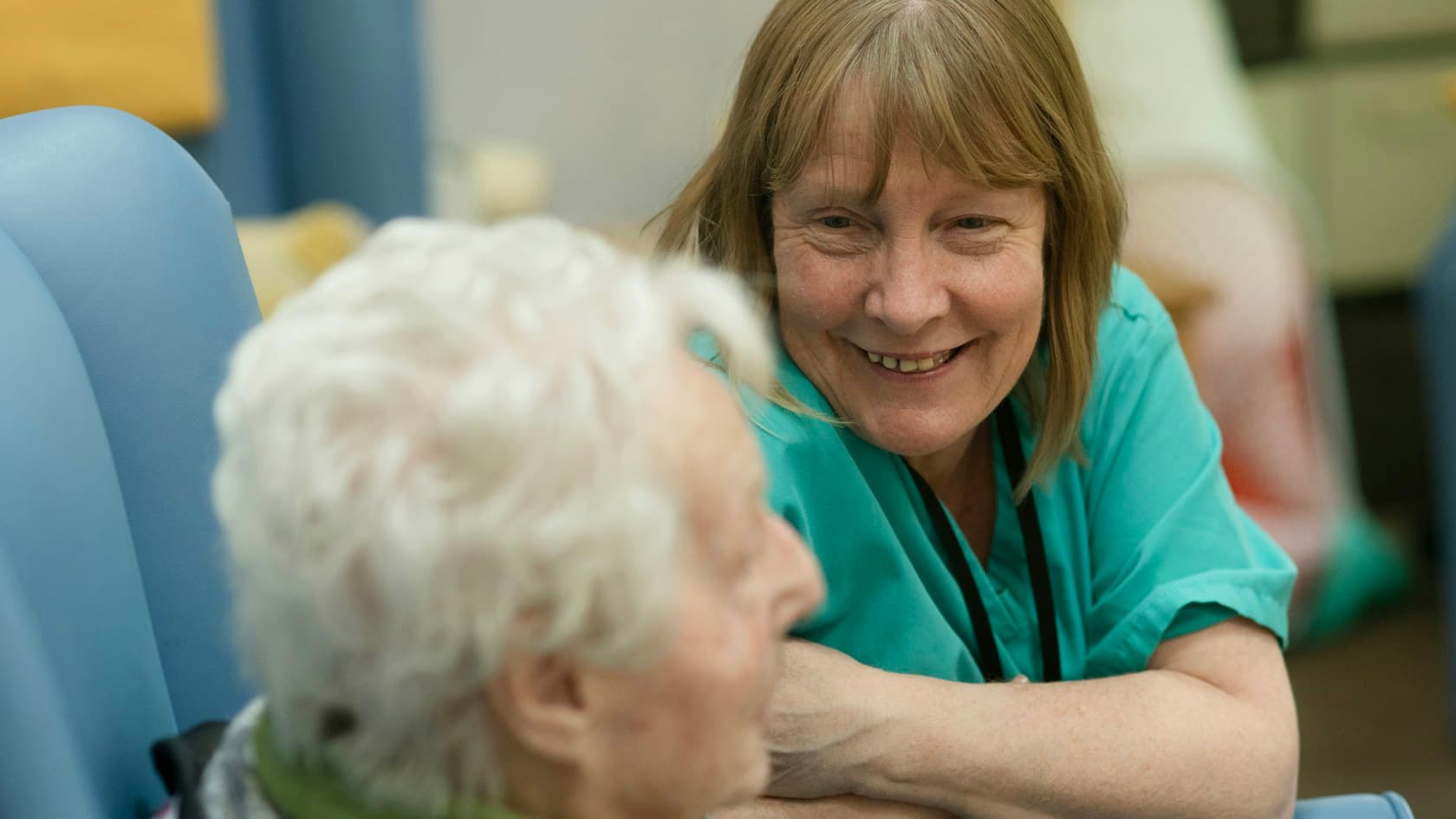 Nurse in green scrubs smiling with an elderly resident in a care home lounge